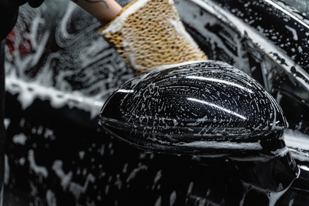 heros-img Hand scrubbing a car mirror with a sponge covered in soap at a car wash.